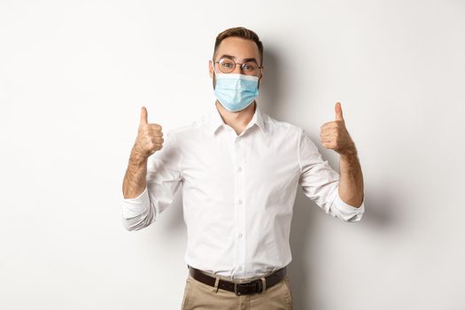 Covid-19, social distancing and quarantine concept. Satisfied male manager showing thumbs up, recommending to wear face mask, standing over white background.