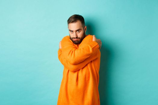 Timid man feeling offended and defensive, hugging himself and looking suspicious at camera, standing over light blue background.