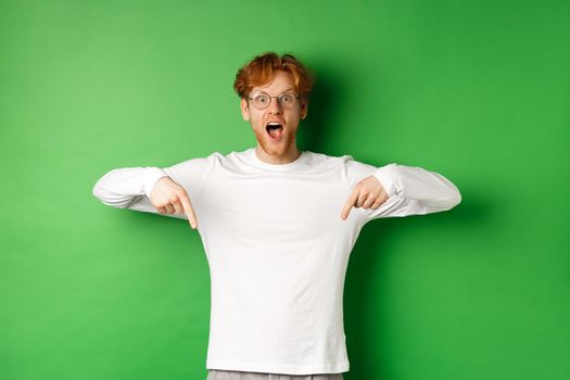 Surprised and excited young redhead man scream of joy and amazement, checking out awesome promo, pointing fingers down, standing over green background.