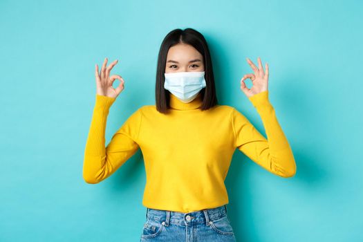 Covid-19, social distancing and pandemic concept. Smiling confident asian woman in medical mask showing OK signs, approve or praise good deal, standing over blue background.