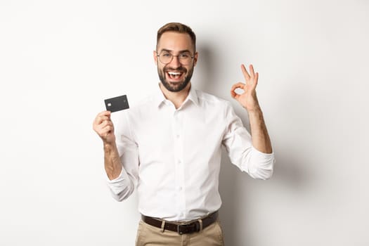 Handsome man showing his credit card and okay sign, recommending bank, standing over white background. Copy space