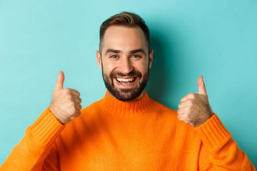 Close-up of attractive bearded man showing thumb-up, approve and like, recommending product, light blue background.