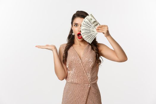 Shopping concept. Shocked woman holding money near face, wearing stylish dress, showing dollars with amazed expression, standing over white background.