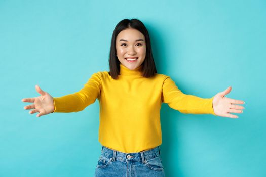 Portrait of beautiful korean woman spread out hands for hug, reaching for cuddles and smiling at camera, greeting you, standing over blue background.