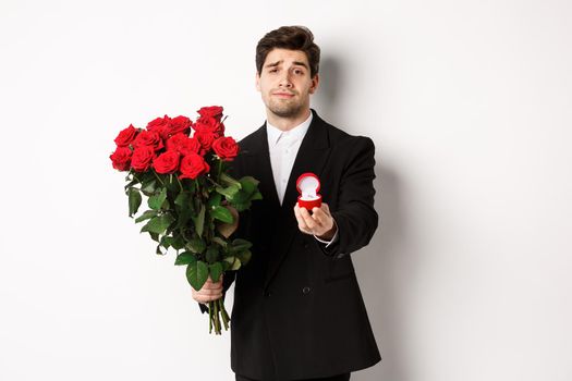 Handsome smiling man in black suit, holding roses and engagement ring, making a proposal to marry him, standing against white background.