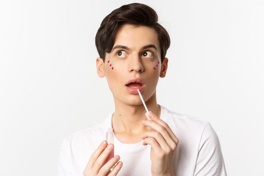 Close-up of beautiful androgynous man with glitter under eyes applying lip gloss, looking dreamy at upper left corner, white background.