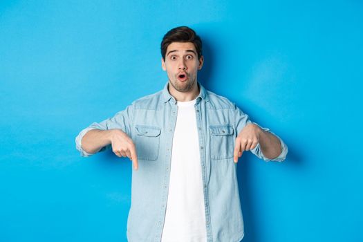 Surprised bearded adult man in casual outfit, pointing fingers down and looking startled, asking question about promotion, standing over blue background.