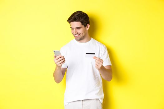 Young man paying online, insert credit card number on mobile phone, shopping in internet, standing over yellow background.