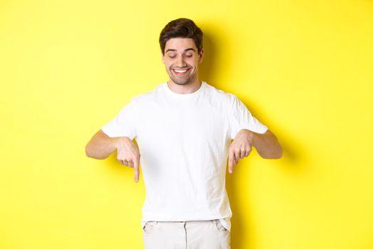 Young handsome man pointing and looking down, checking out promo offer, standing over yellow background.