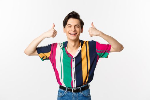 Young cheerful man smiling, showing thumbs up in approval, praise something good, standing over white background.