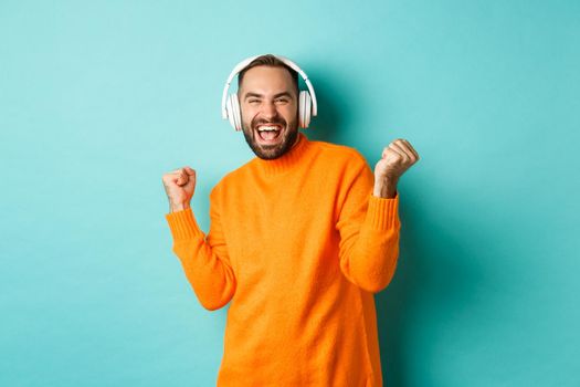 Happy adult man in orange sweater, looking up and listening music in headphones, standing over blue background.