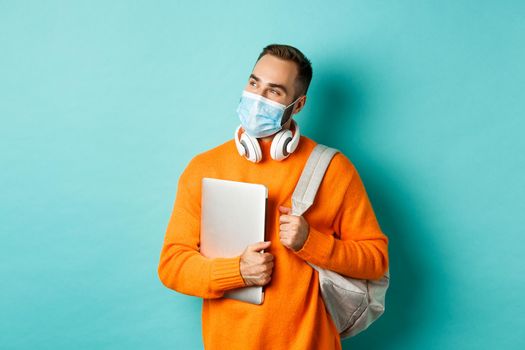 Handsome caucasian man with headphones and backpack, holding laptop and wearing medical mask, looking happy, standing over light blue background.