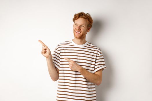 Cheerful caucasian guy with red hair and beard, looking and pointing left at logo banner, standing over white background.