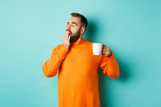 Sleepy handsome man drinking coffee and yawning, standing in orange sweater against light blue background.