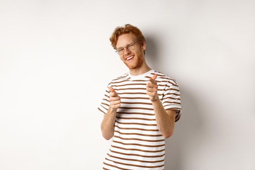 Cool and sassy redhead man with beard and glasses, pointing finger pistols at camera and smiling, congratulating you, standing over white background.