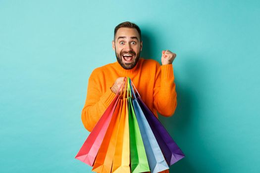 Lifestyle concept. Excited man showing shopping bags and rejoicing from discounts, standing over turquoise background.