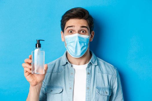 Concept of covid-19, pandemic and quarantine. Surprised guy in medical mask holding hand sanitizer bottle, raising eyebrows amazed, standing over blue background.