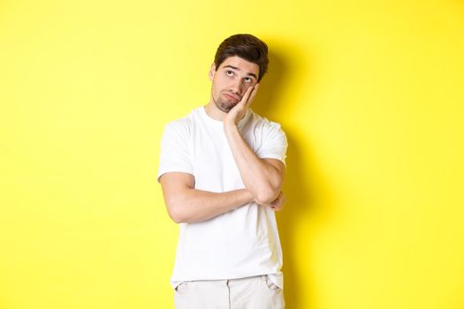 Bored and gloomy guy looking up, imaging things, standing over yellow background.