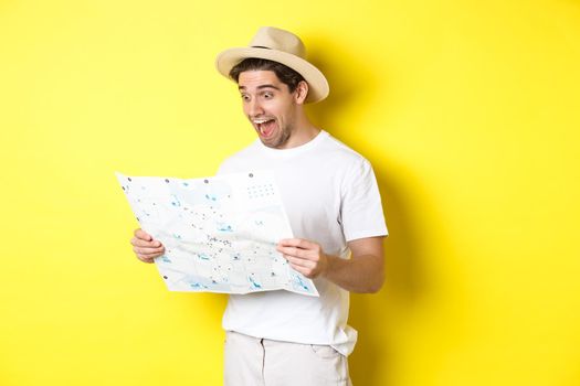 Travelling, vacation and tourism concept. Smiling happy tourist looking at map with sightseeings, standing against yellow background.