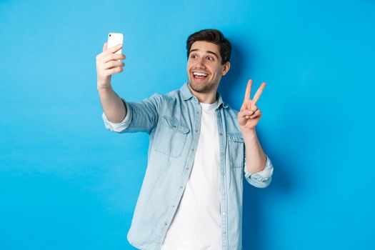 Happy man taking selfie and showing peace sign on blue background, holding mobile phone.