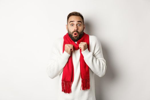 Christmas holidays and celebration concept. Man looking in awe at camera, standing surprised, feeling cold on new year, wearing red scarf and sweater, white background.