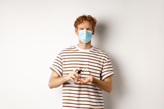 Covid, virus and social distancing concept. Teenager redhead guy in face mask clean hands with sanitizer, using antiseptic, standing over white background.