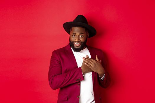 Fashion and party concept. Handsome african american macho man flirting, smiling and holding hands on heart, being grateful and touched, standing over red background.