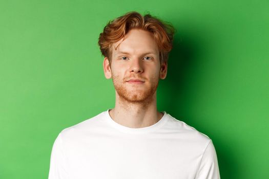 Close-up of young man with red messy hair and beard looking at camera, standing over green background.