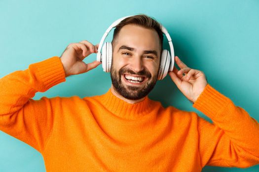 Close-up of handsome modern man listening music in headphones, standing in orange sweater over turquoise background.