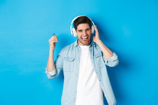 Happy smiling man enjoying listening to music in headphones, holding smartphone in raised hand, standing over blue background.