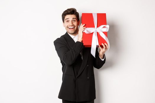 Concept of christmas holidays, celebration and lifestyle. Attractive man in black suit, holding new year gift and smiling, standing with a present over white background.