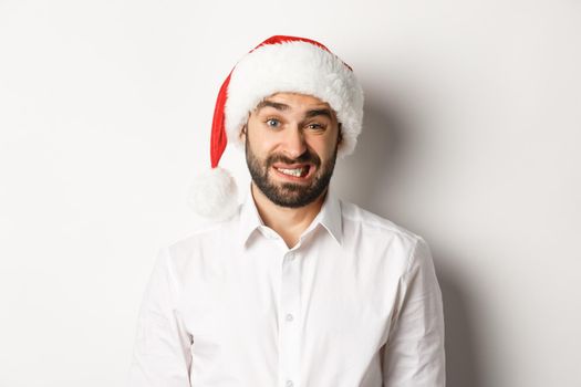 Close-up of awkward guy in santa hat apologizing, feeling uncomfortable, standing over white background. Christmas concept.