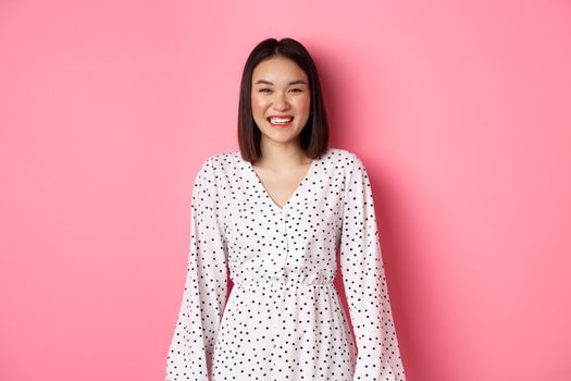 Happy korean woman in dress looking at camera, smiling and laughing with sincere expression, standing over pink background. Copy space