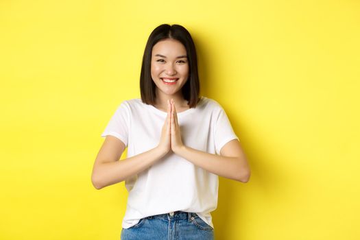 Beautiful asian woman say thank you, holding hands in namaste, pray gesture and smiling, being grateful, standing over yellow background.