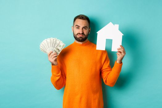 Real estate concept and mortgage concept. Sad man showing house maket and money, looking upset and disappointed, standing over light blue background.