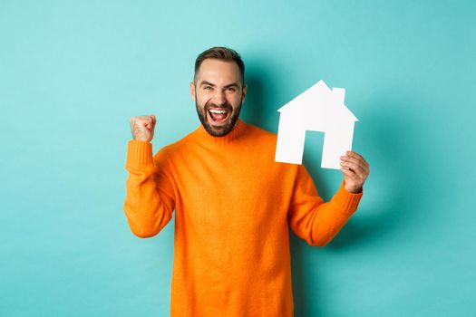 Real estate concept. Excited man saying yes, showing paper house maket and looking satisfied, standing in orange sweater over light blue background.