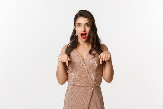 Amazed beautiful woman in glamour dress celebrating christmas party, pointing fingers down at promo, standing over white background.