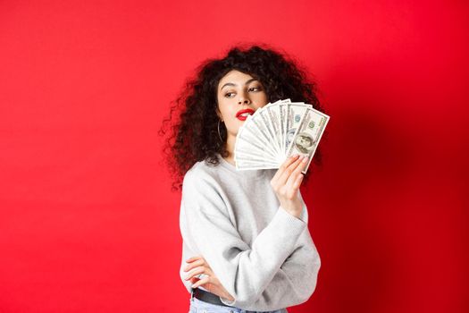 Beautiful rich woman looking sensual aside, waving at herself with dollar bills fan, standing seductive on red background.