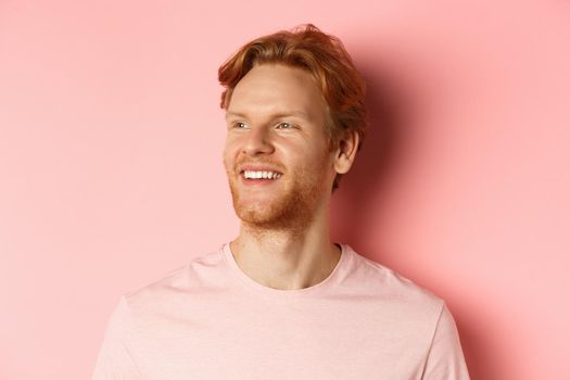 Close up of handsome young man with beard and red hair, looking left and smiling delighted, standing confident over pink background.