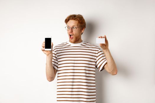 Shopping and finance concept. Amazed young man with red hair showing plastic credit card and smartphone blank screen, staring at display impressed, white background.