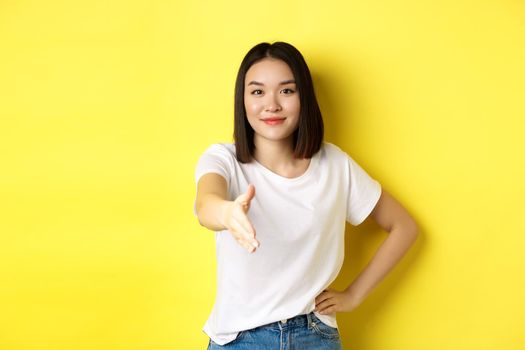 Confident asian woman in white t-shirt, stretch out hand for handshake and greeting gesture, saying hello, introduce herself, standing over yellow background.