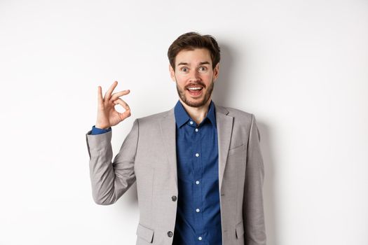 Excited bearded guy in business suit gasping amazed, looking with admiration and showing okay sign, say yes, approve and like good product, standing on white background.