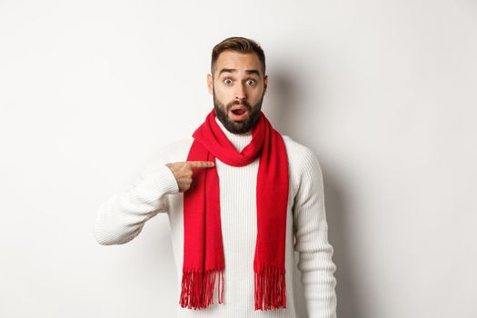 Winter holidays and shopping concept. Surprised and confused guy pointing at himself, being chosen, standing in red scarf and sweater against white background.