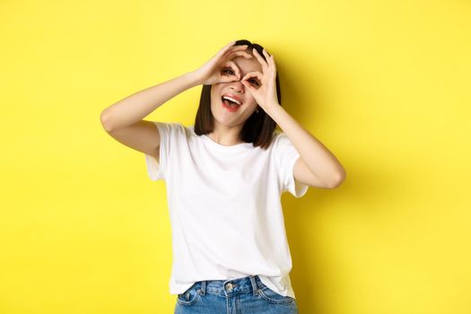 Funny asian girl looking through hand binoculars and smiling, stading over yellow background.