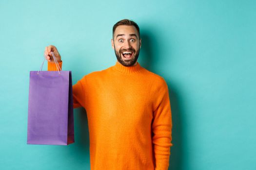 Excited adult man holding purple shopping bag and smiling, going to mall, standing over turquoise background.