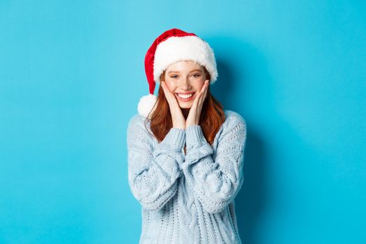 Winter holidays and Christmas Eve concept. Cheerful redhead girl in santa hat, celebrating New Year, blushing and smiling happy, standing over blue background.