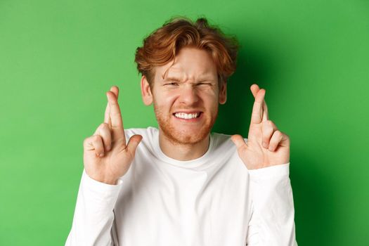 Hopeful caucasian man looking worried, waiting for important results and making wish with fingers crossed, standing over green background.