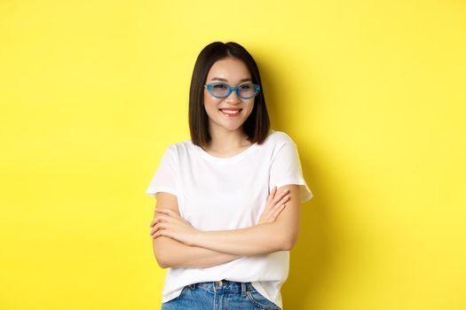 Fashion and lifestyle concept. Sassy and confident asian woman in trendy sunglasses looking self-assured at camera, standing over yellow background.