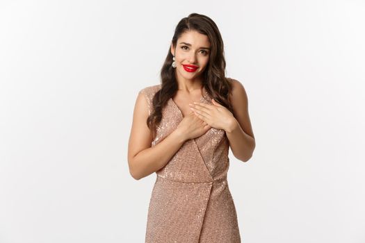 Image of beautiful young woman staring grateful at camera, holding hands on heart touched and thankful, receiving gift, standing in evening dress at party, white background.