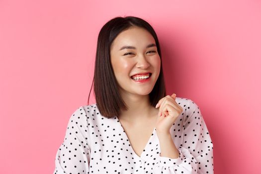 Beauty and lifestyle concept. Close-up of happy asian female laughing, looking happy and showing genuine emotions, standing over pink background.